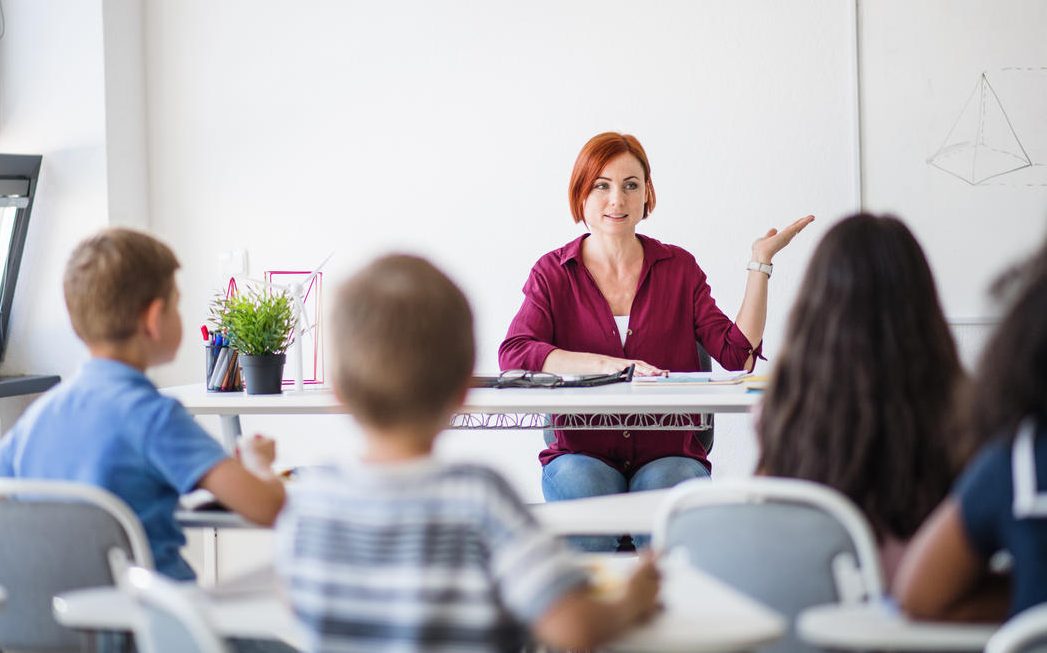 Rear view of school children sitting at the desk in classroom, listening to teacher.